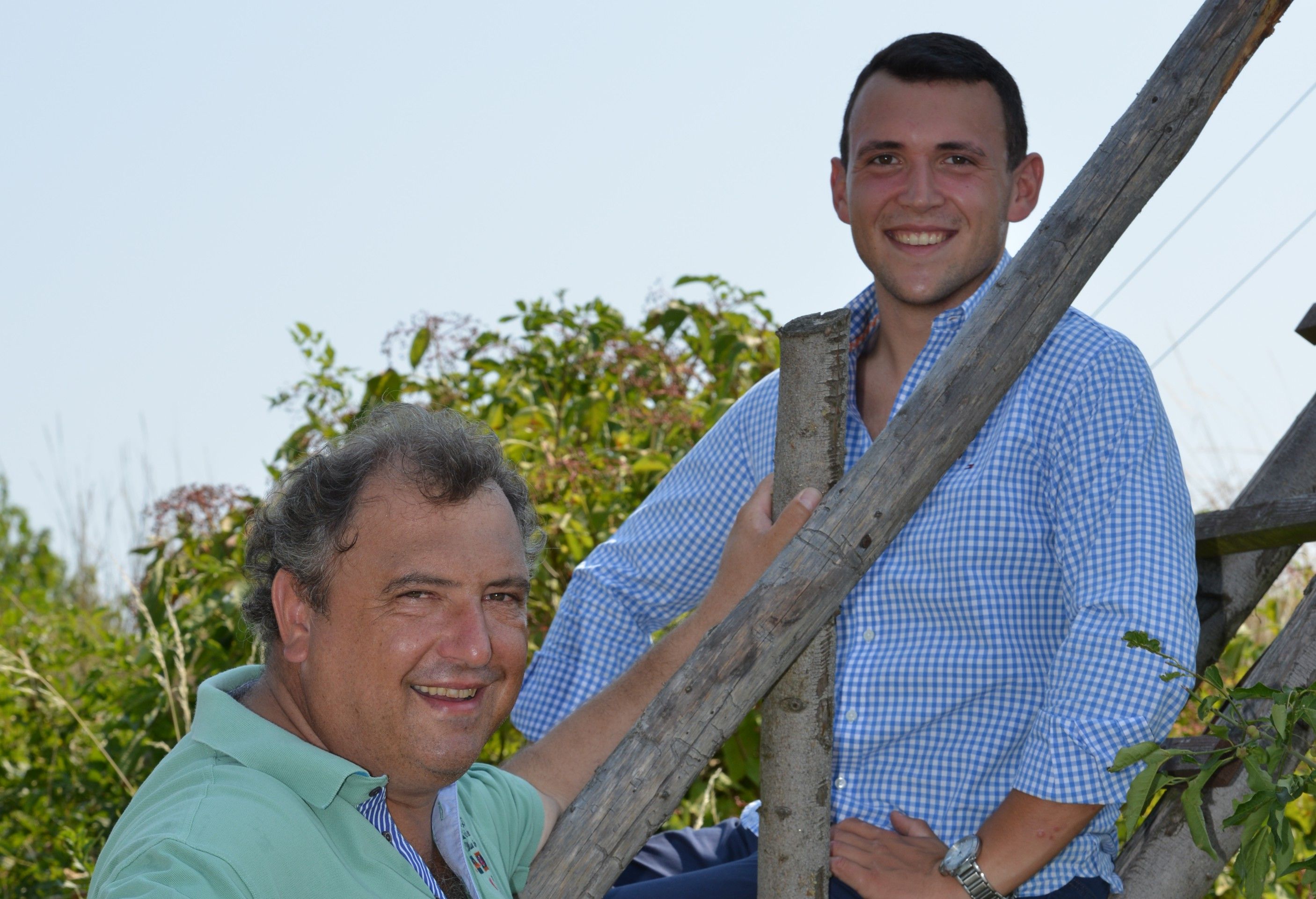 Zwei Männer lächeln in die Kamera, einer sitzt, der andere steht auf einer Holzleiter im Freien.