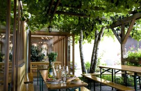 Gem&uuml;tliche Terrasse mit Holztischen, Weinflasche und Gl&auml;sern, umgeben von gr&uuml;nen Pflanzen und Weinreben.