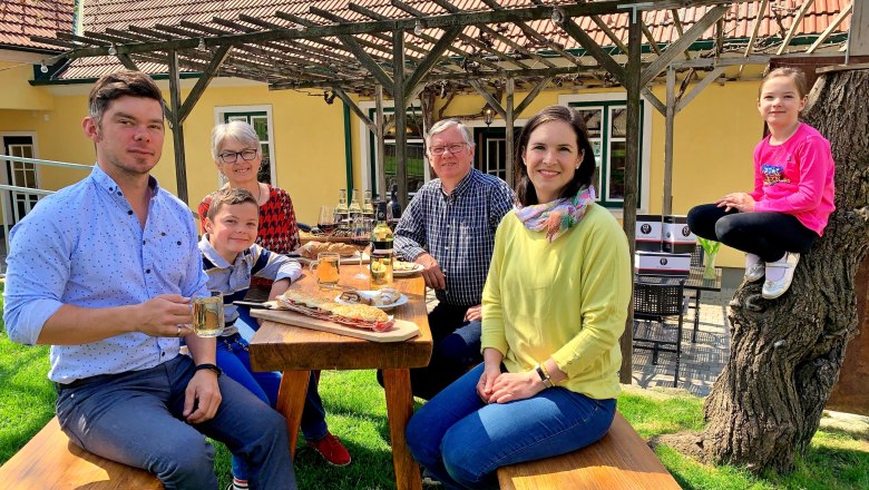 Eine Familie sitzt an einem Holztisch im Freien und genießt eine Mahlzeit. Ein Kind sitzt auf einem Baumstamm. Im Hintergrund ein gelbes Haus mit Pergola.
