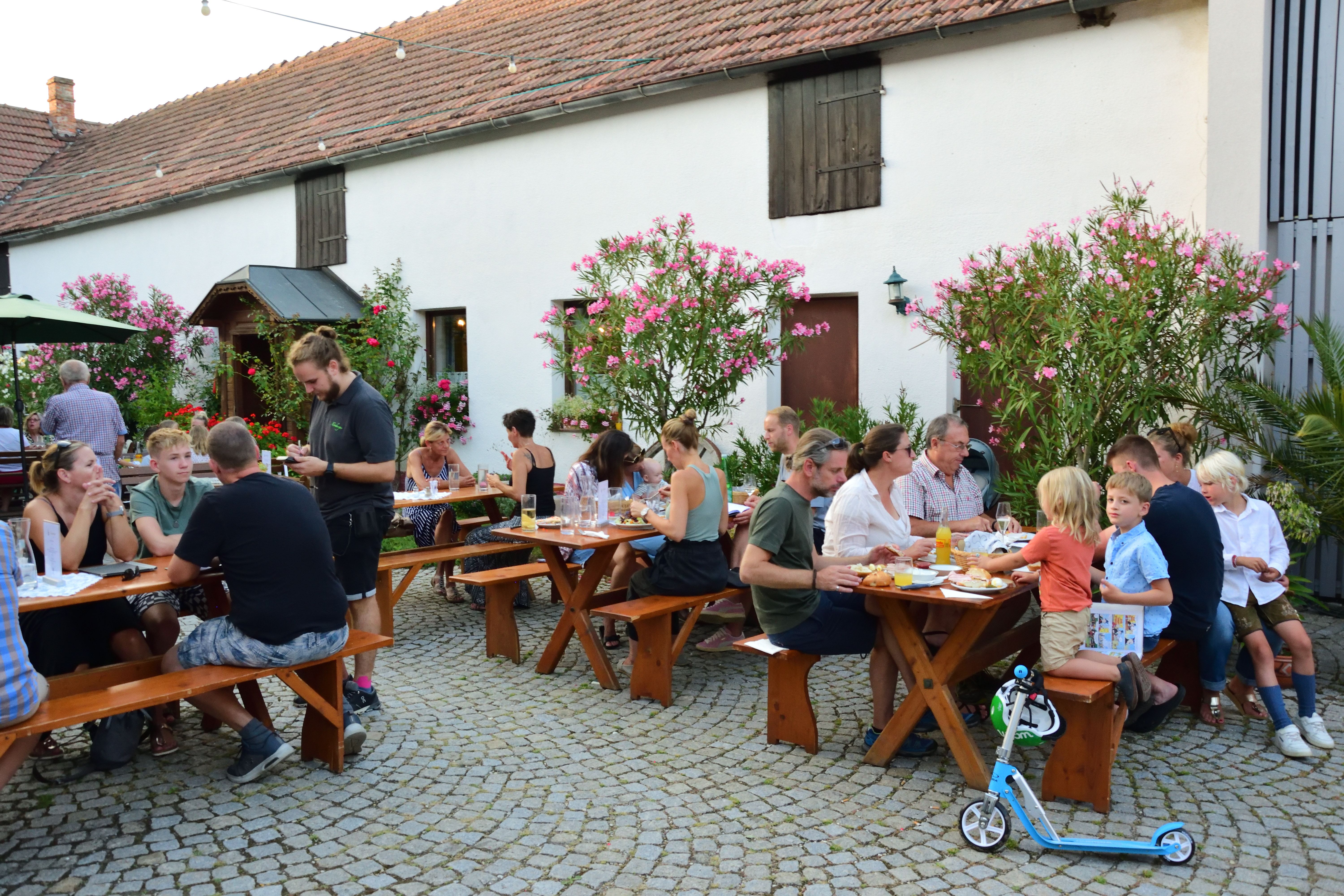 Menschen sitzen an Holztischen im Freien eines Gastgartens, umgeben von blühenden Sträuchern.