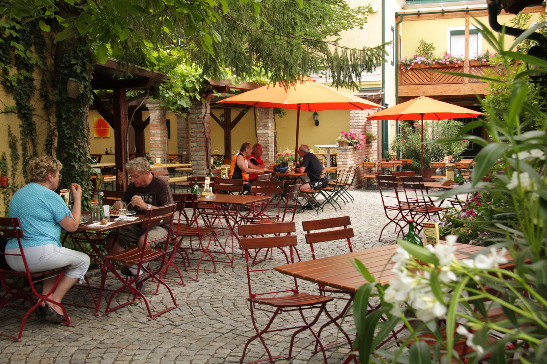 Ein gemütlicher Biergarten mit Holztischen, roten Stühlen und orangefarbenen Sonnenschirmen. Menschen sitzen und essen im Freien.