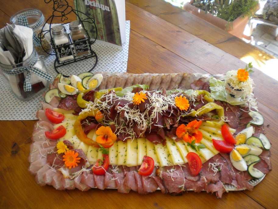 Eine reichhaltige Heurigenplatte mit verschiedenen Wurst- und Käsesorten, dekoriert mit Gemüse und essbaren Blüten auf einem Holztisch.