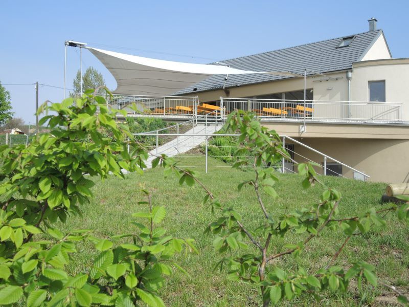 Heurigenlokal mit Terrasse und Sonnensegel, umgeben von grüner Landschaft.
