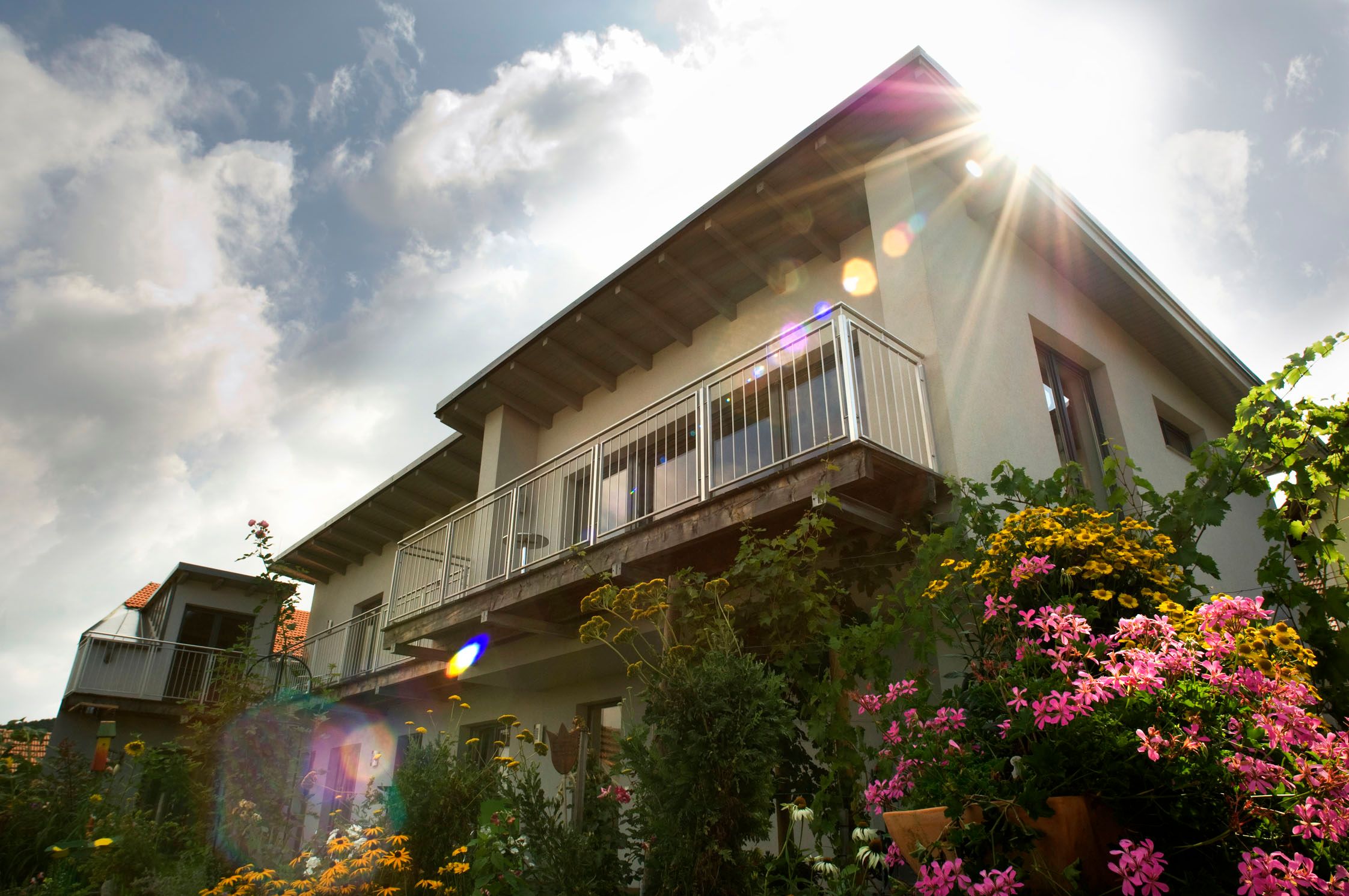 Außenansicht eines Hauses mit Balkon und blühendem Garten im Vordergrund, Sonnenstrahlen am Himmel.