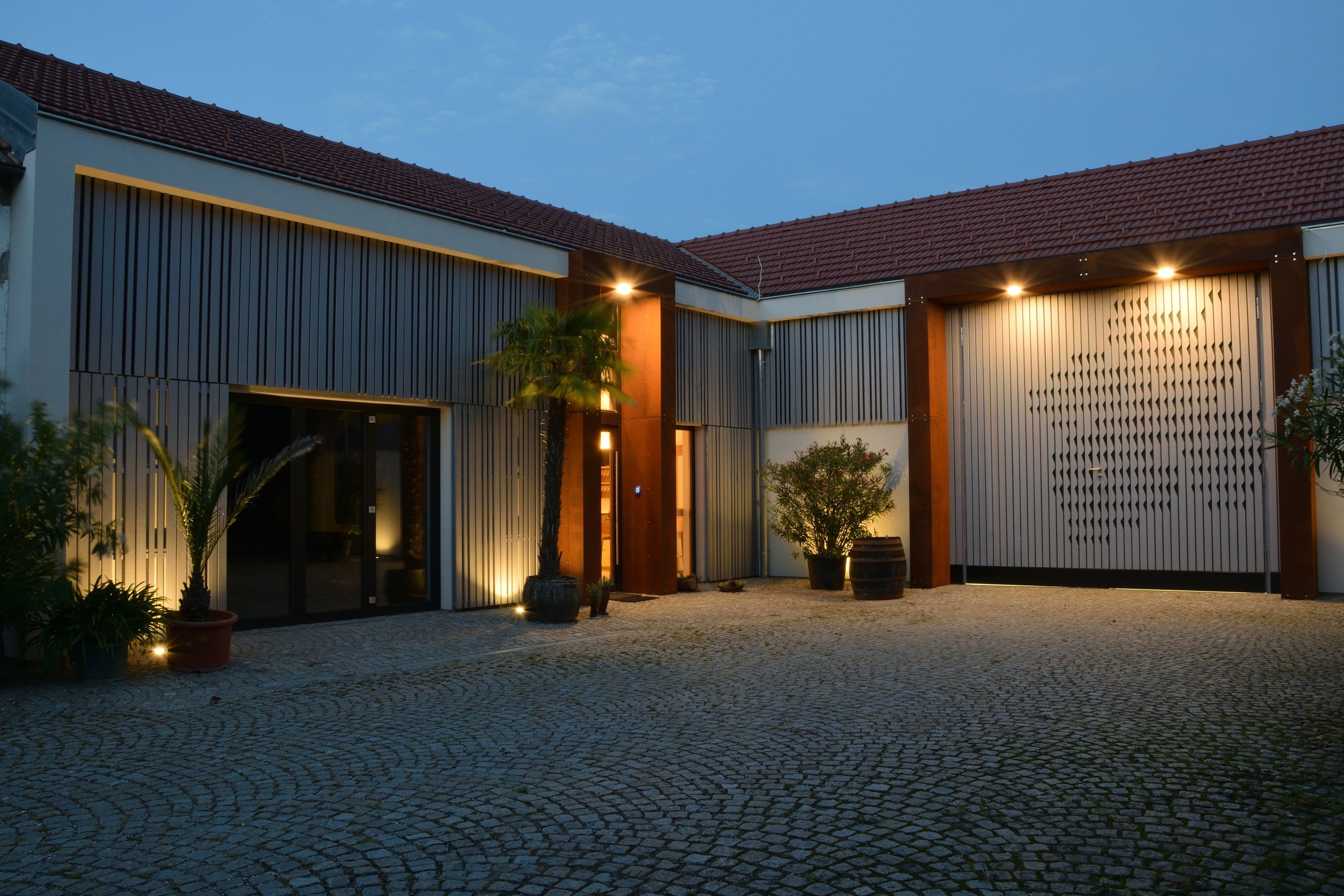 Beleuchteter Innenhof einer modernen Kellerei mit Palmen und Pflastersteinen.