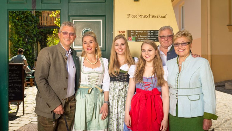 Eine Familie in traditioneller Tracht vor einem Geb&auml;ude mit der Aufschrift 'Flaschenweinverkauf'.