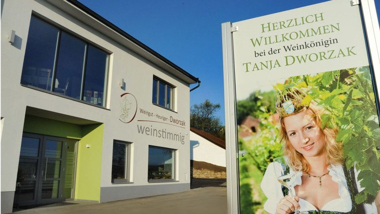 Ein modernes Gebäude mit einem Schild, das eine Frau in Tracht mit einem Weinglas zeigt. Text: Herzlich willkommen bei der Weinkönigin Tanja Dworzak.