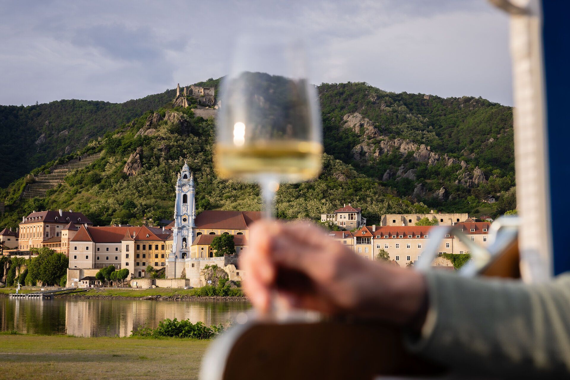 Jemand hält ein Weißweinglas in die Kamera und im Hintergrund ist Dürnstein zu sehen.