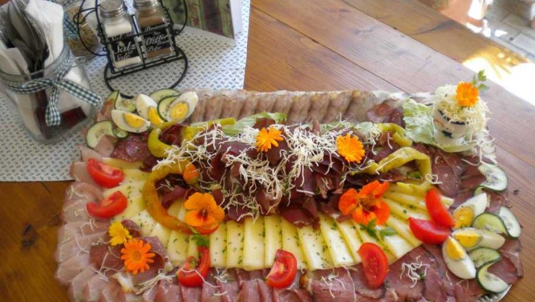 Eine reichhaltige Heurigenplatte mit verschiedenen Wurst- und K&auml;sesorten, dekoriert mit Gem&uuml;se und essbaren Bl&uuml;ten auf einem Holztisch.