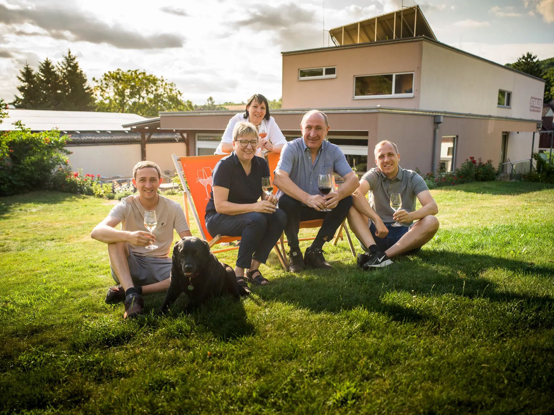 Eine Familie sitzt mit einem Hund auf einer Wiese vor einem modernen Haus und hält Weingläser.