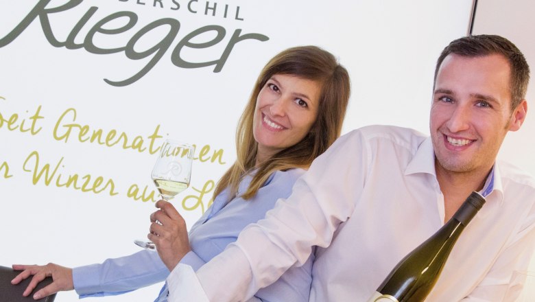 Zwei Personen lächeln und halten Weinflaschen vor einem Schild mit der Aufschrift 'Oberschil Rieger'.