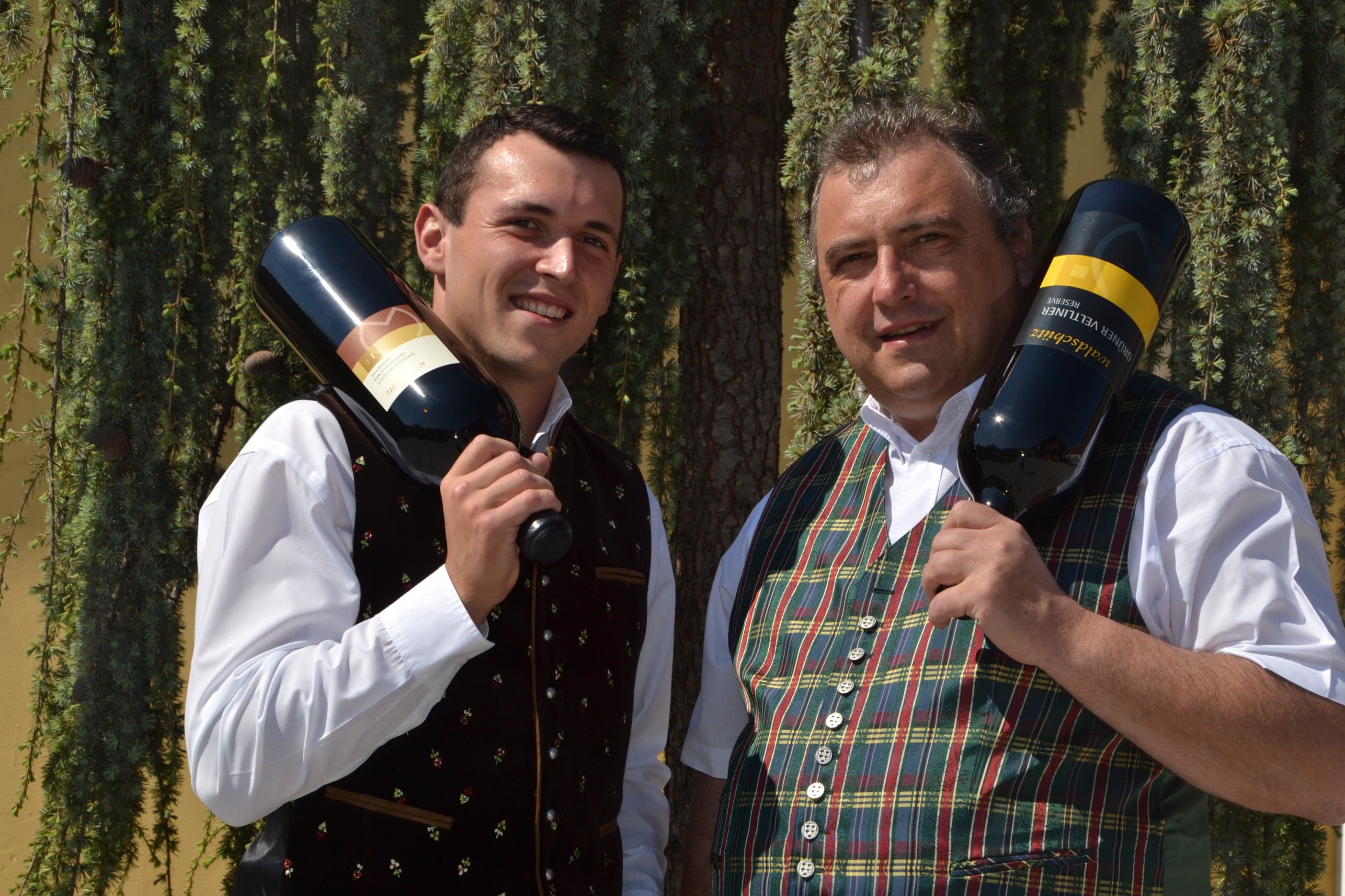 Zwei Männer in traditioneller Kleidung halten große Weinflaschen vor einem Baum.