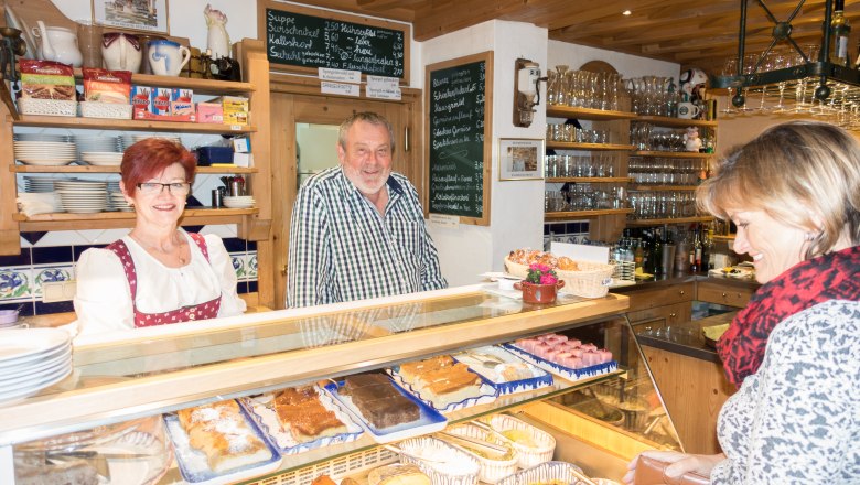 Ein Mann und eine Frau stehen hinter einer Theke in einem Caf&eacute;, w&auml;hrend eine Kundin vorne steht. Auf der Theke sind verschiedene Kuchen ausgestellt.