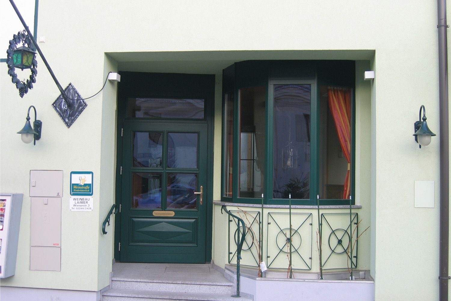 Eingangsbereich eines Gebäudes mit grüner Tür und Fenstern, dekoriert mit Pflanzen und Laternen.